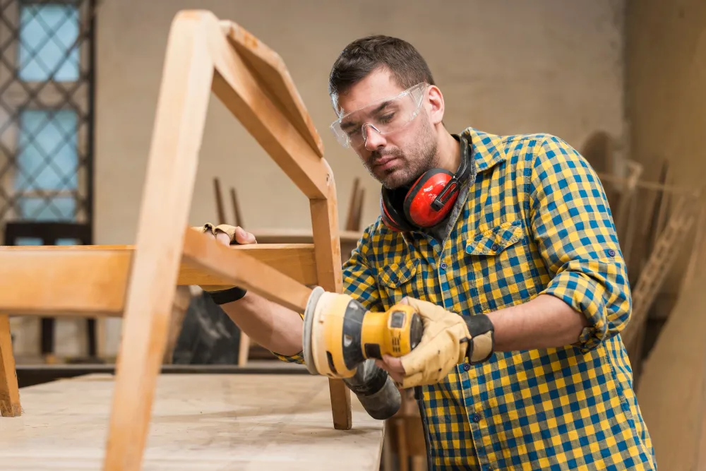 A male carpenter wearing safety glasses using sander on furniture in workshop